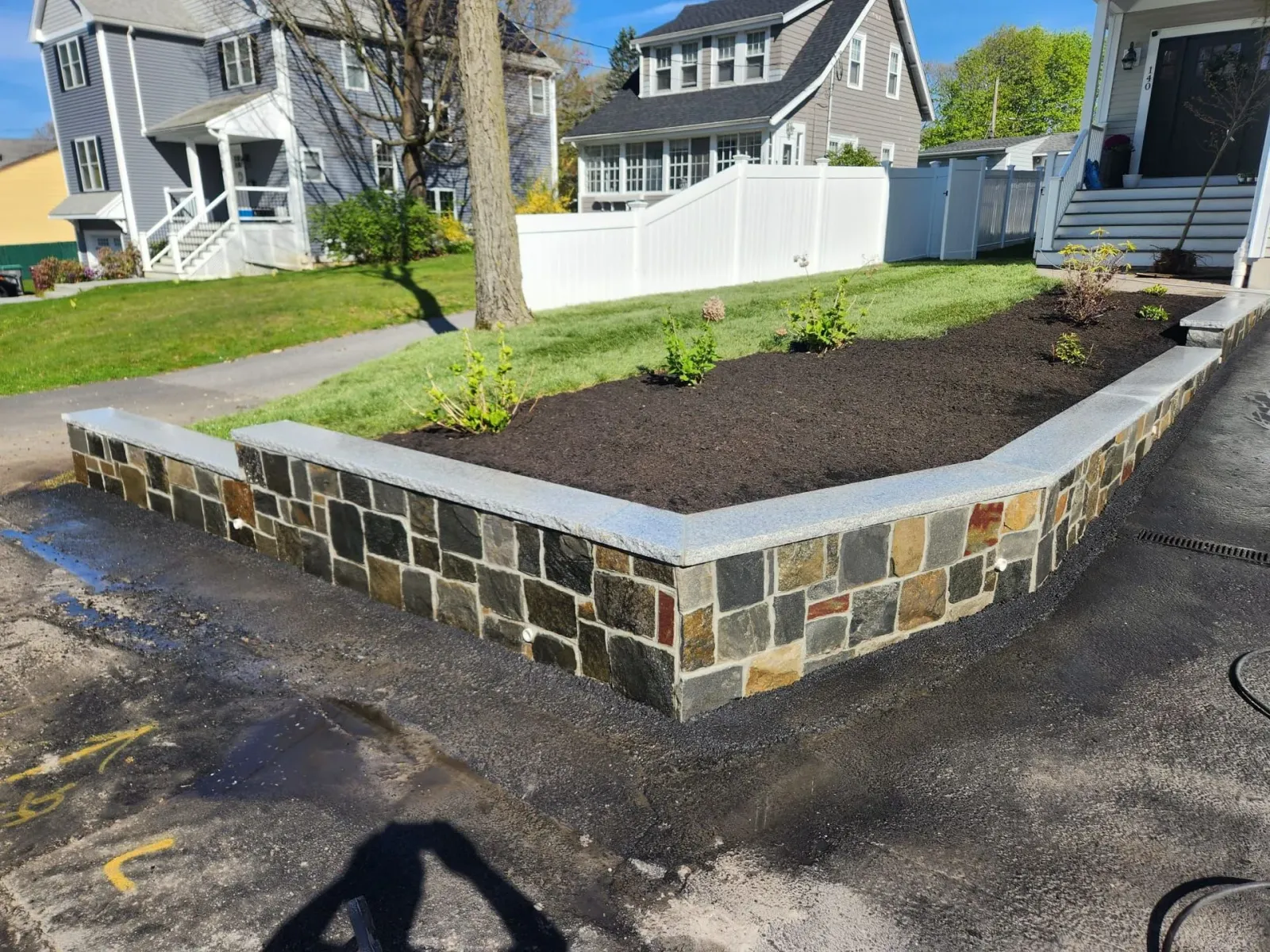 Raised stone planter with dark mulch and a small tree beside a white fence in a suburban yard