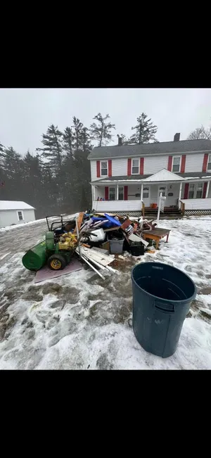 A large pile of debris and trash sits on snow-covered ground in front of a white two-story house with red shutters.