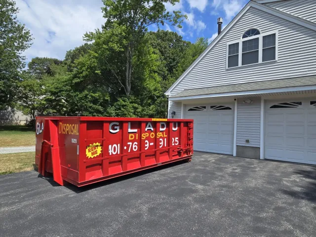 Red dumpster with yellow and white lettering in driveway next to a white house with garage doors. Trees and blue sky in the background.