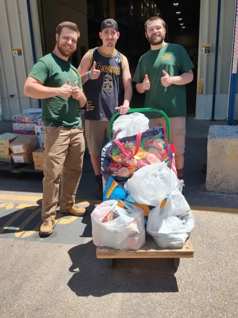Three volunteers in green shirts stand behind a cart loaded with bags of donations.