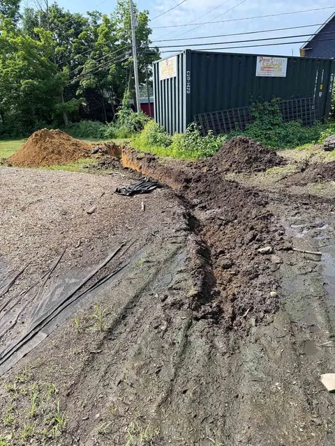 Mud-filled dirt road beside a green shipping container and piles of soil under bright sun