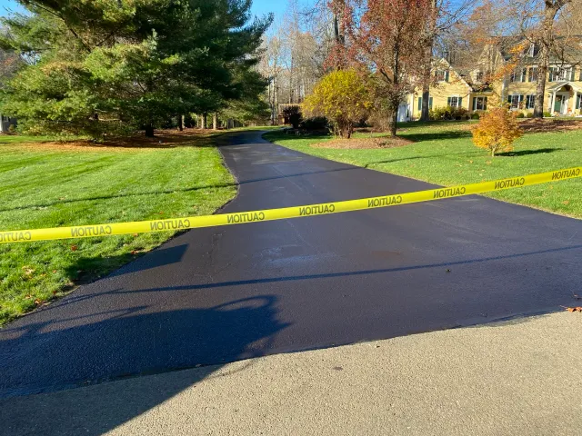 A residential street with a freshly paved black asphalt road, blocked off by yellow caution tape reading 'CAUTION' in black, and houses with trees in the background.