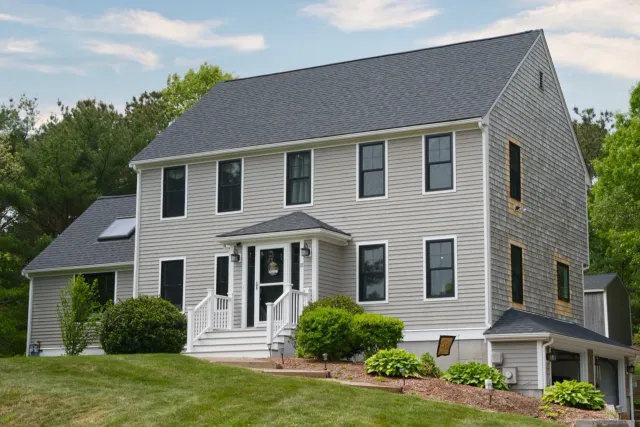 A three-story suburban house with beige siding, black-framed windows, and a dark gray roof, surrounded by a well-maintained lawn and green trees.