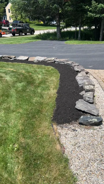 A landscaped yard with a stone border, mulch bed, and green lawn, next to a gravel path and driveway.