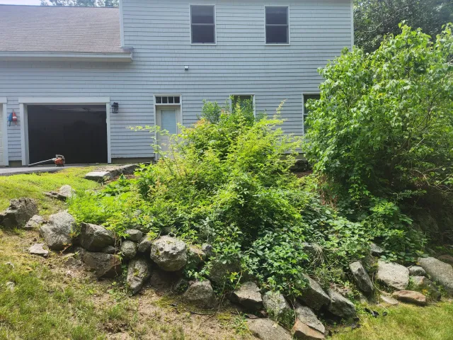A backyard with a stone retaining wall, green bushes, and a house with a garage door, two windows, and white siding.