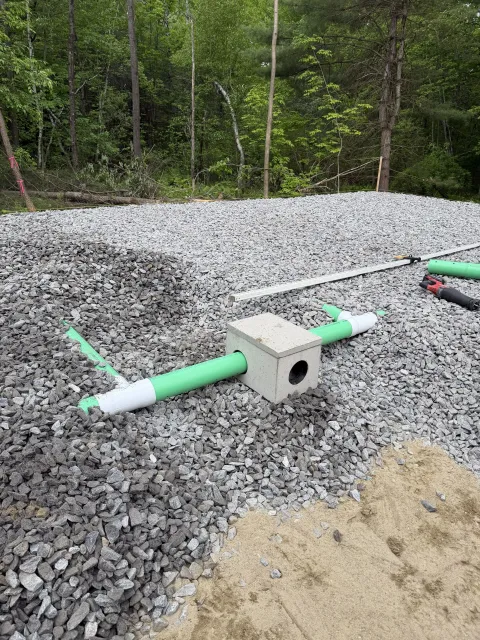 Gravel drainage bed with green PVC pipes and a concrete block, set beside a wooded area