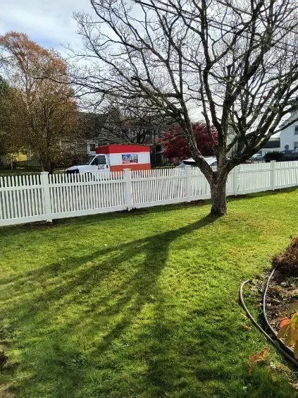 White picket fence with a tree casting a shadow on green grass, white moving truck in the background.