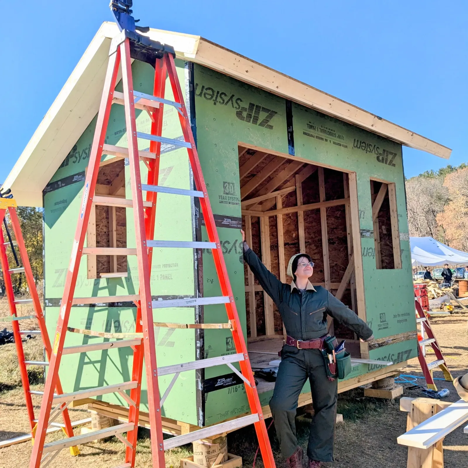 A person wearing safety glasses and work clothes stands in front of a small house under construction, smiling and pointing upward, with ladders nearby and construction materials around.