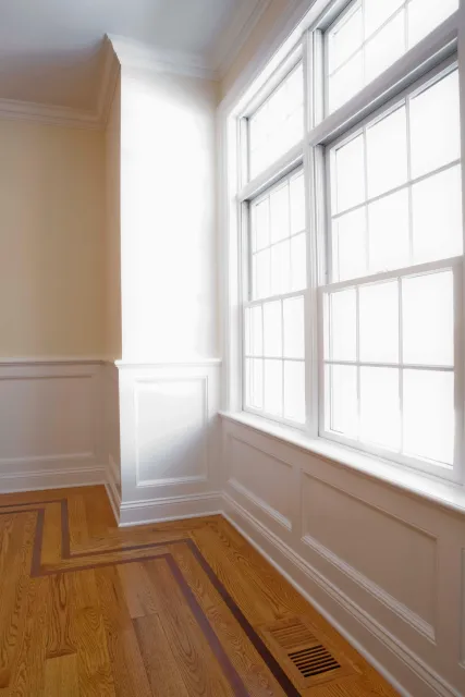 Bright corner of a room with large windows and light wood flooring. The walls are painted white with decorative panels and a vent on the floor. Sunlight is streaming through the windows.