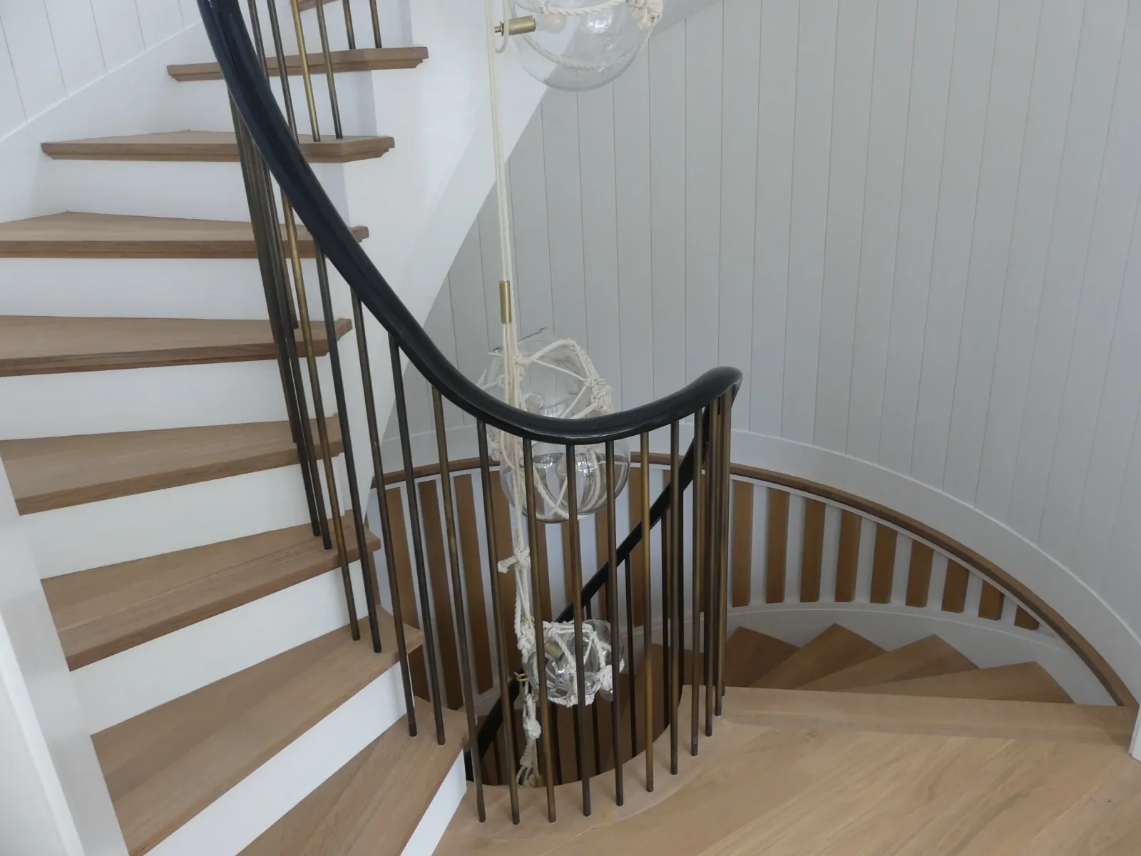 Wooden staircase with black and brass railing, spiral down in a home interior with white paneled walls, and a glass and rope hanging light fixture.
