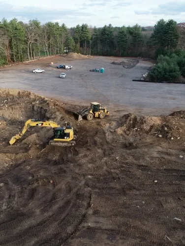 An aerial view shows excavators and loaders performing earthwork at a large cleared construction site surrounded by trees.
