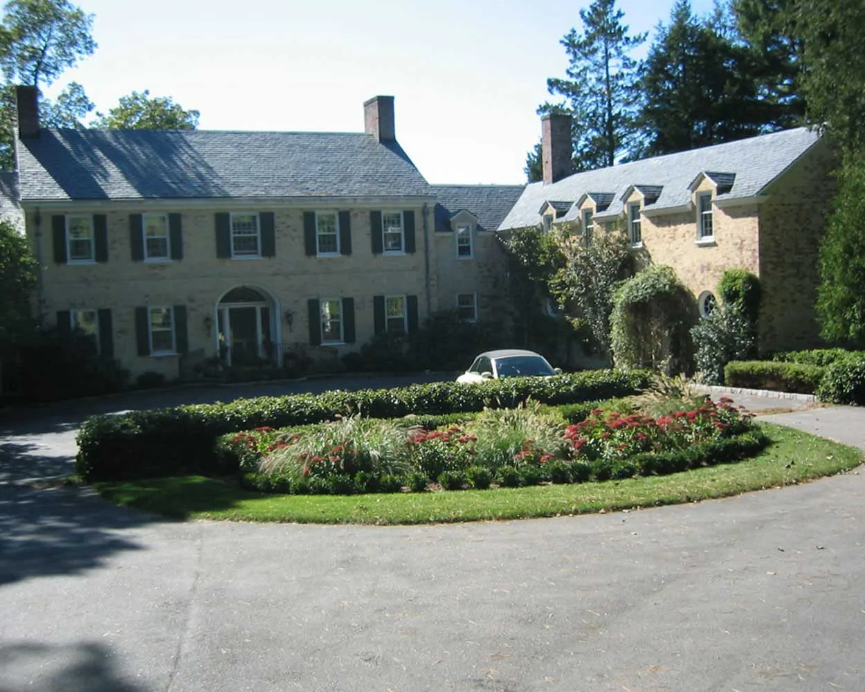 Large brick colonial home with black shutters, surrounded by lush landscaping and a circular driveway featuring a central garden bed filled with ornamental grasses and red flowering plants.