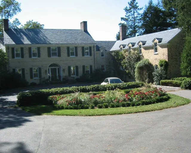 Large brick colonial home with black shutters, surrounded by lush landscaping and a circular driveway featuring a central garden bed filled with ornamental grasses and red flowering plants.
