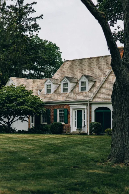 Cypress Works Massachusetts, a front view of a two-story house with a brick exterior, green shutters, and dormer windows on the roof, surrounded by trees and a well-maintained lawn.