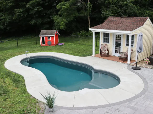 A backyard with a kidney-shaped fiberglass swimming pool, a small yellow house with a porch and chairs, a patio umbrella, a black fence, and a red doghouse with a gray door on a grassy lawn surrounded by trees.