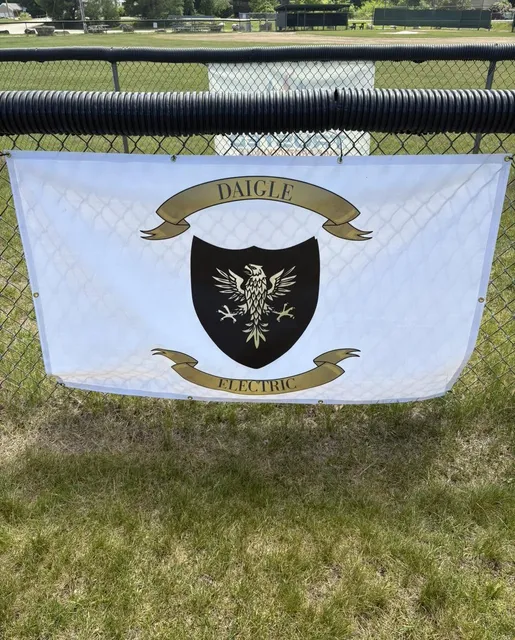 The Daigle Electric banner — white with the black eagle shield — hanging on the outfield fence at the Barrington ballfields.