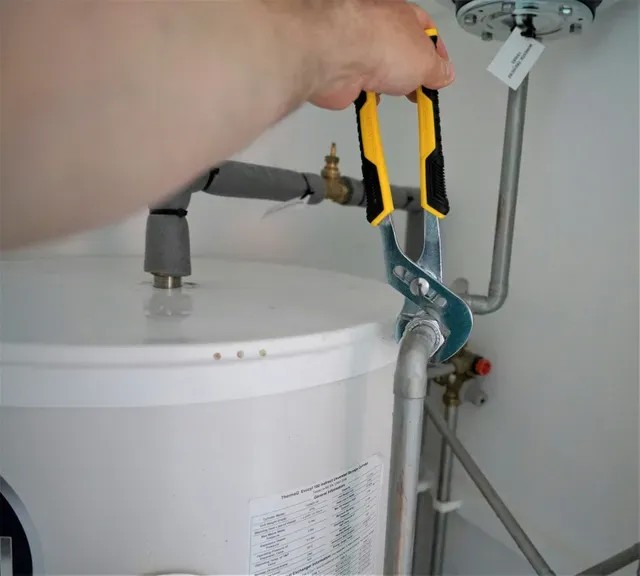 A person using large pliers to adjust a metal pipe connected to the top of a white water heater tank.