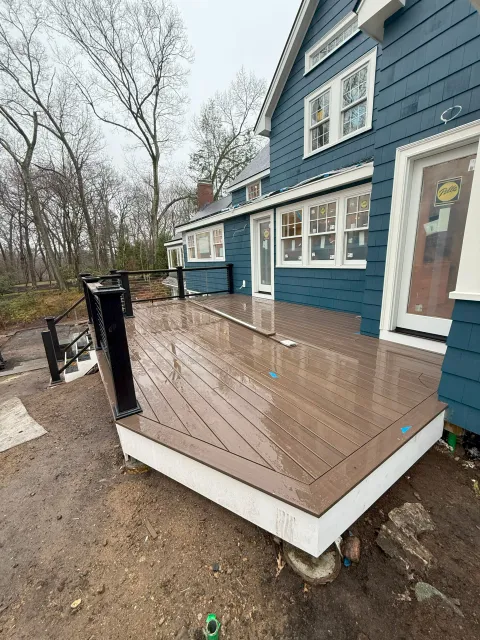 Newly built brown composite deck with black posts and cable railing attached to a dark blue house under construction.