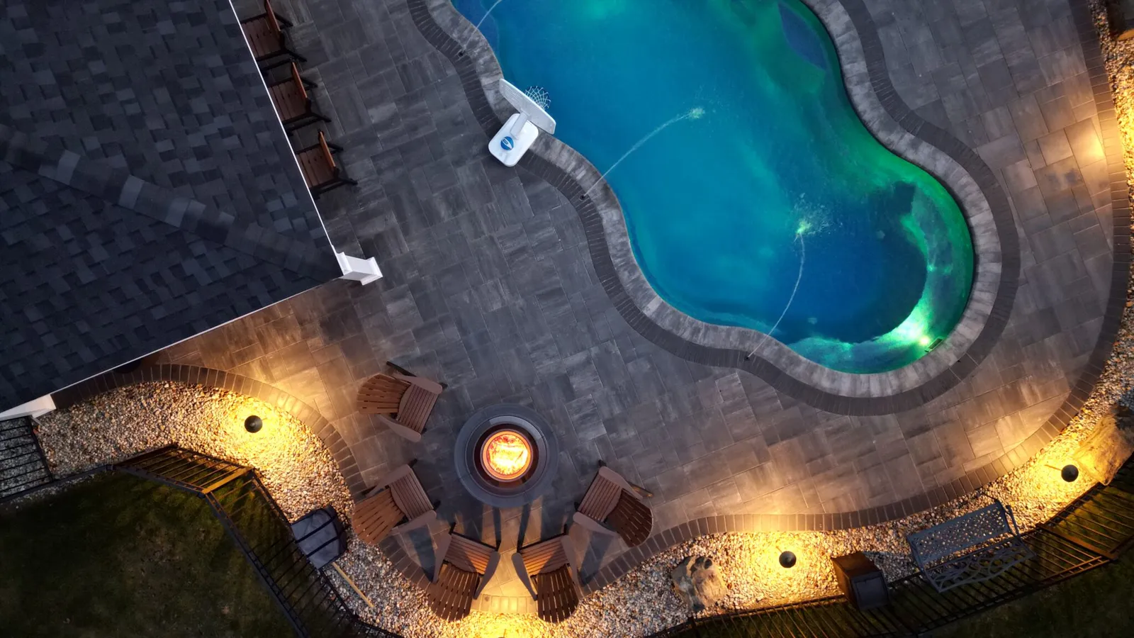 Top-down aerial view of a backyard featuring a freeform swimming pool with underwater lighting, a circular fire pit surrounded by Adirondack chairs, curved stone paver patio, and landscape lighting along the perimeter.