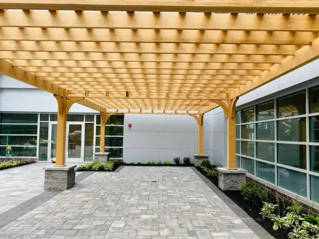 Wooden pergola over a gray brick patio, beside a building with large windows and a white wall.