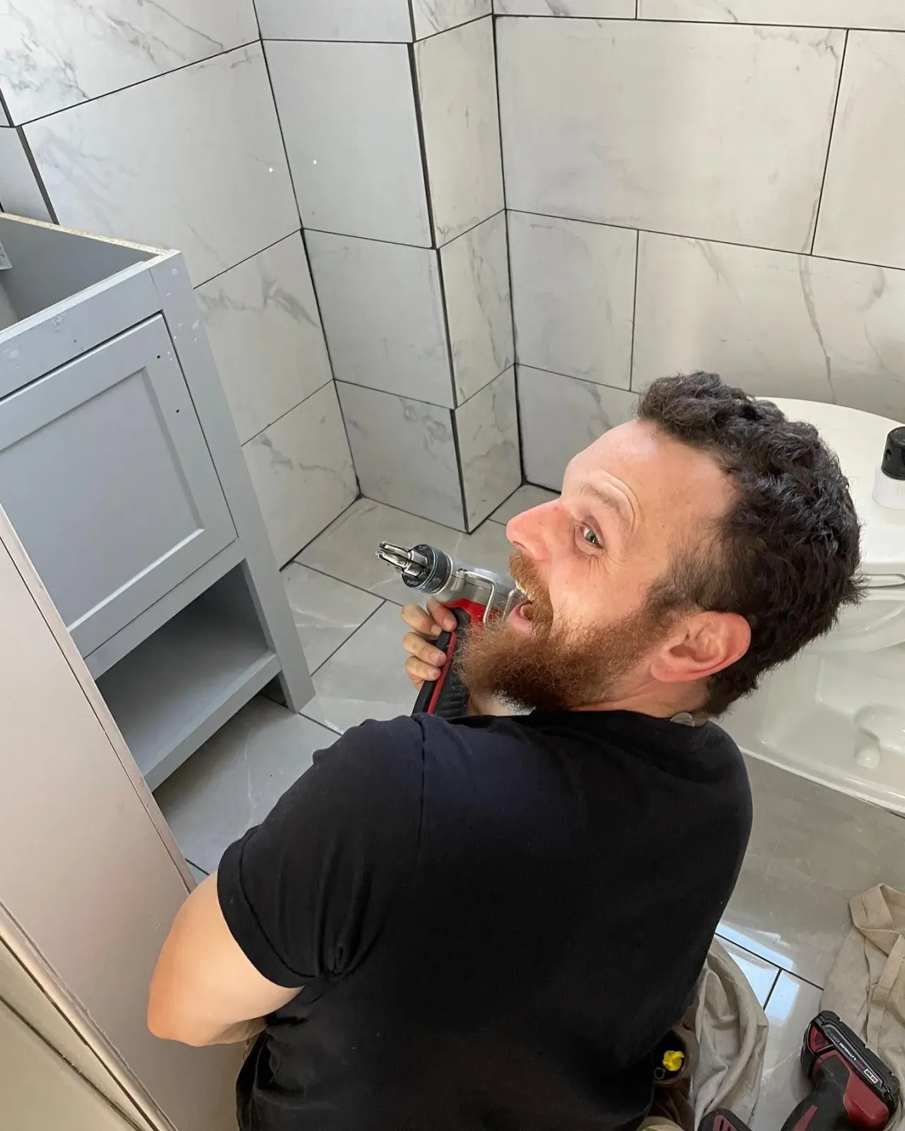 A man with a beard is using a drill in a bathroom.