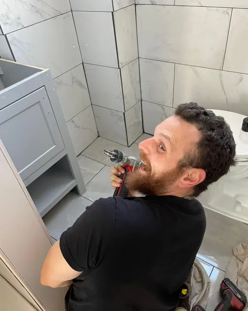 A man with a beard is using a drill in a bathroom.