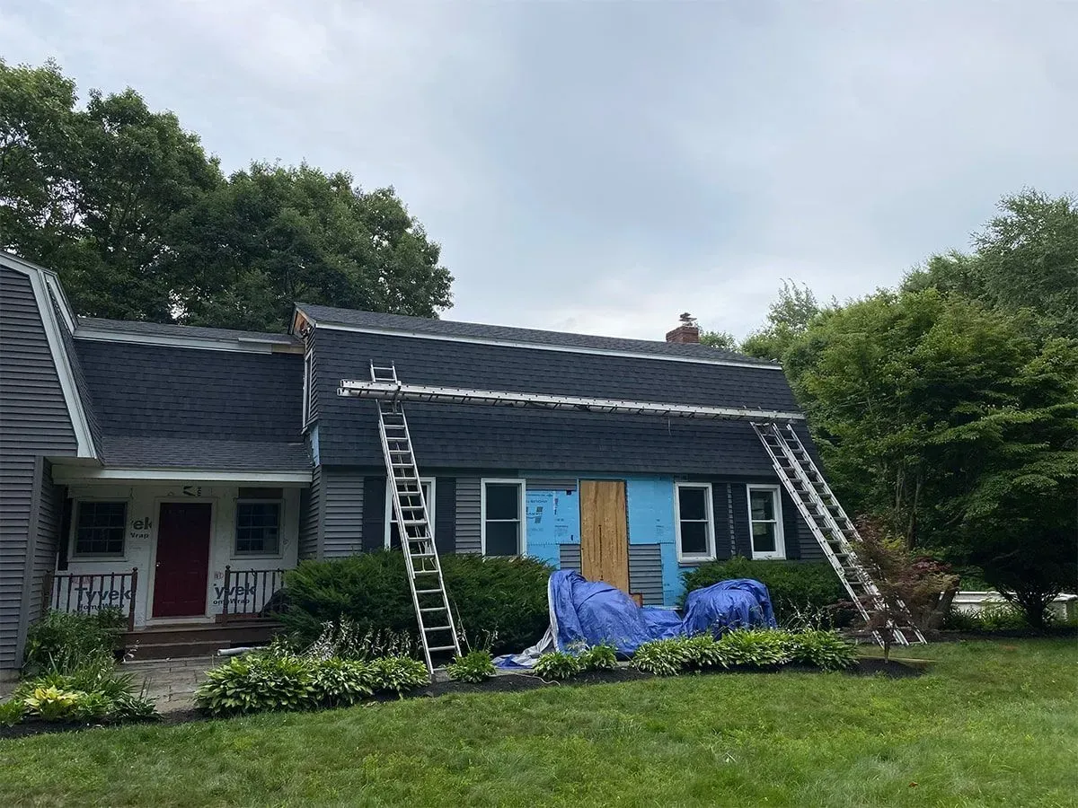 Two-story house with blue siding and ladders leaning against the roof, under repair after storm damage