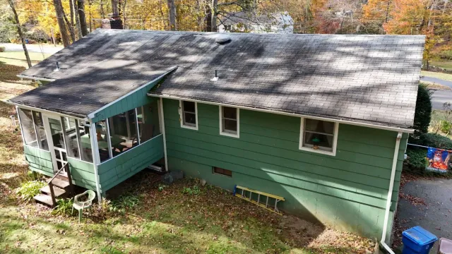 Green house with attached screened porch and sloping roof; fall foliage in the background.