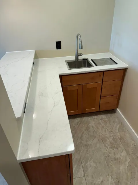 L-shaped countertop with sink, faucet, and wooden cabinets in a small kitchen area with light tan walls.