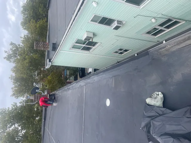 Person in red working on a flat roof with tools, near a chimney and satellite dish, next to a building with green siding and windows.
