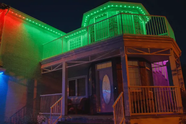 House illuminated with permanent lighting in vibrant green, red, blue, and purple. The colorful lights accent the roofline and porch of the two-story home at night.