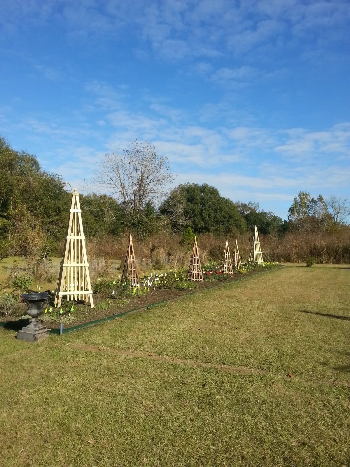Cypress Works Massachusetts, a French Villa in Louisiana featuring a garden with several wooden plant supports, green grass, and trees in the background under a blue sky with scattered clouds.