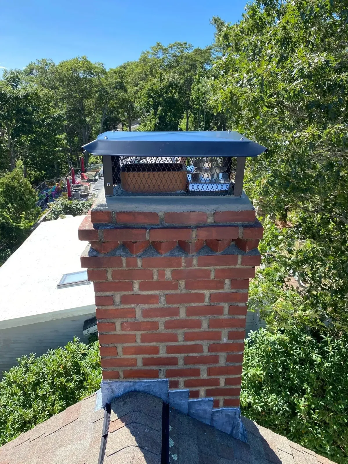 A brick chimney on a shingled roof, topped with a black metal cap, surrounded by green trees on a sunny day.
