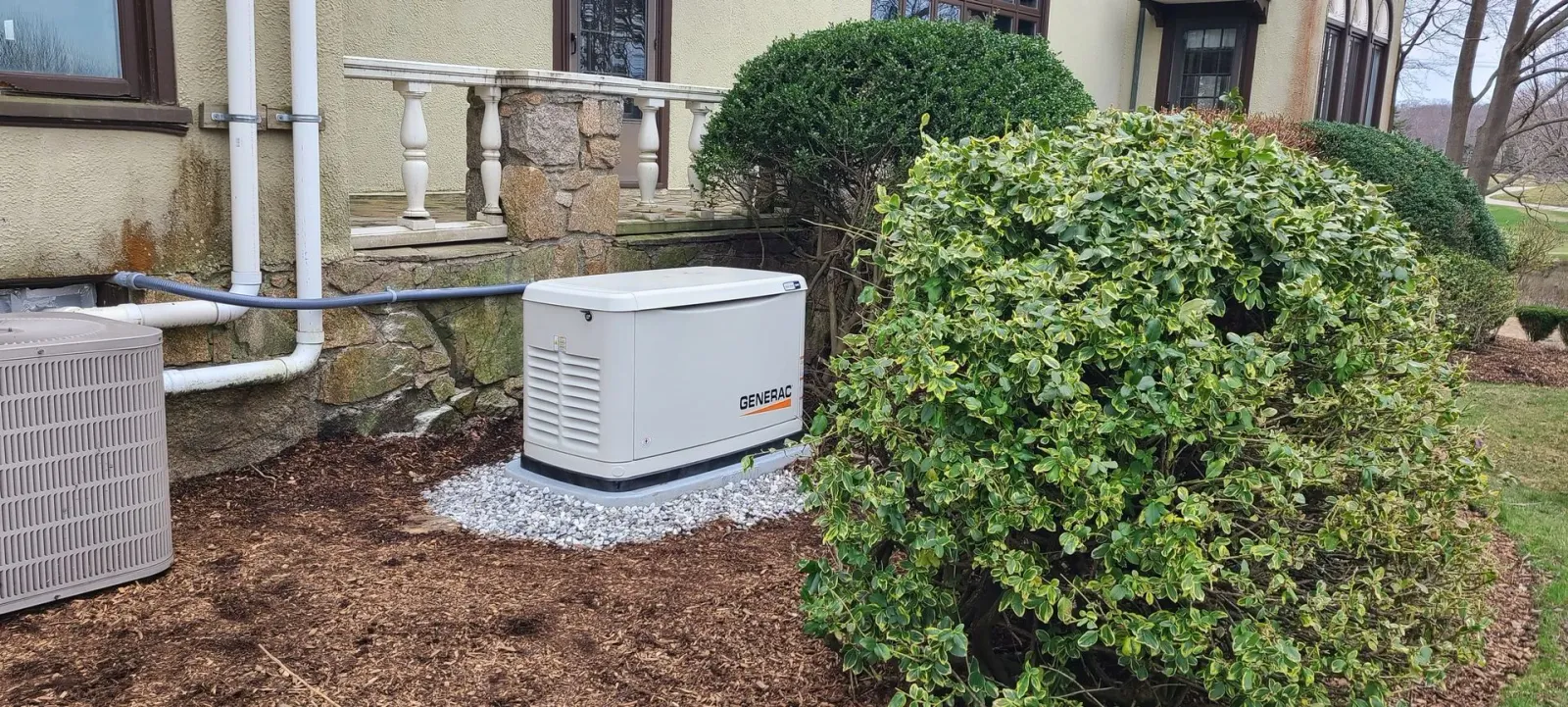 Outdoor HVAC unit beside a house, partly hidden by a large green shrub and mulch bed