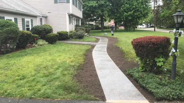 Newly laid concrete walkway curving through a green yard with bushes, trees, and a house in the background.