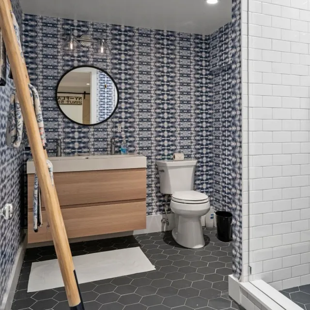 Modern bathroom with blue patterned wallpaper, a round mirror, a wooden vanity with two sinks, a toilet, a black trash bin, and a white tiled shower wall. There is a white bath mat on the hexagonal black tile floor.