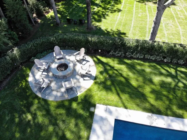 Aerial view of a backyard with a circular fire pit surrounded by eight Adirondack chairs, green grass, trees casting shadows, a hedge, a patio, and a swimming pool.