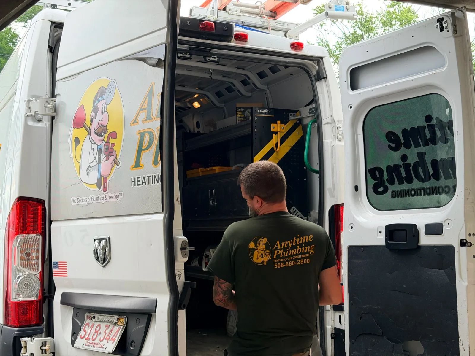 A worker from Anytime Plumbing, wearing a black shirt with yellow lettering, is inside a white plumbing service van. The van has a graphic of a cartoon plumber character and the company's contact information. The worker is facing away and appears to be preparing tools or equipment from the van.
