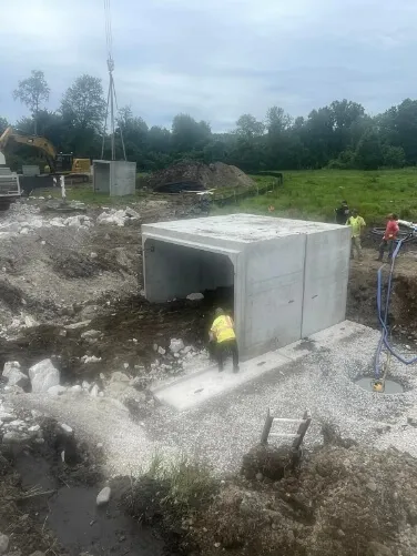 Workers install a precast concrete box culvert at an active construction site using heavy machinery and excavation equipment.