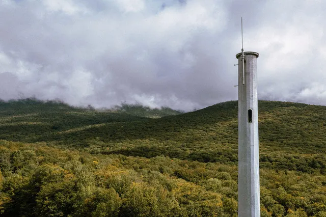 A tall white cylindrical tower stands in a forested valley under cloudy skies.