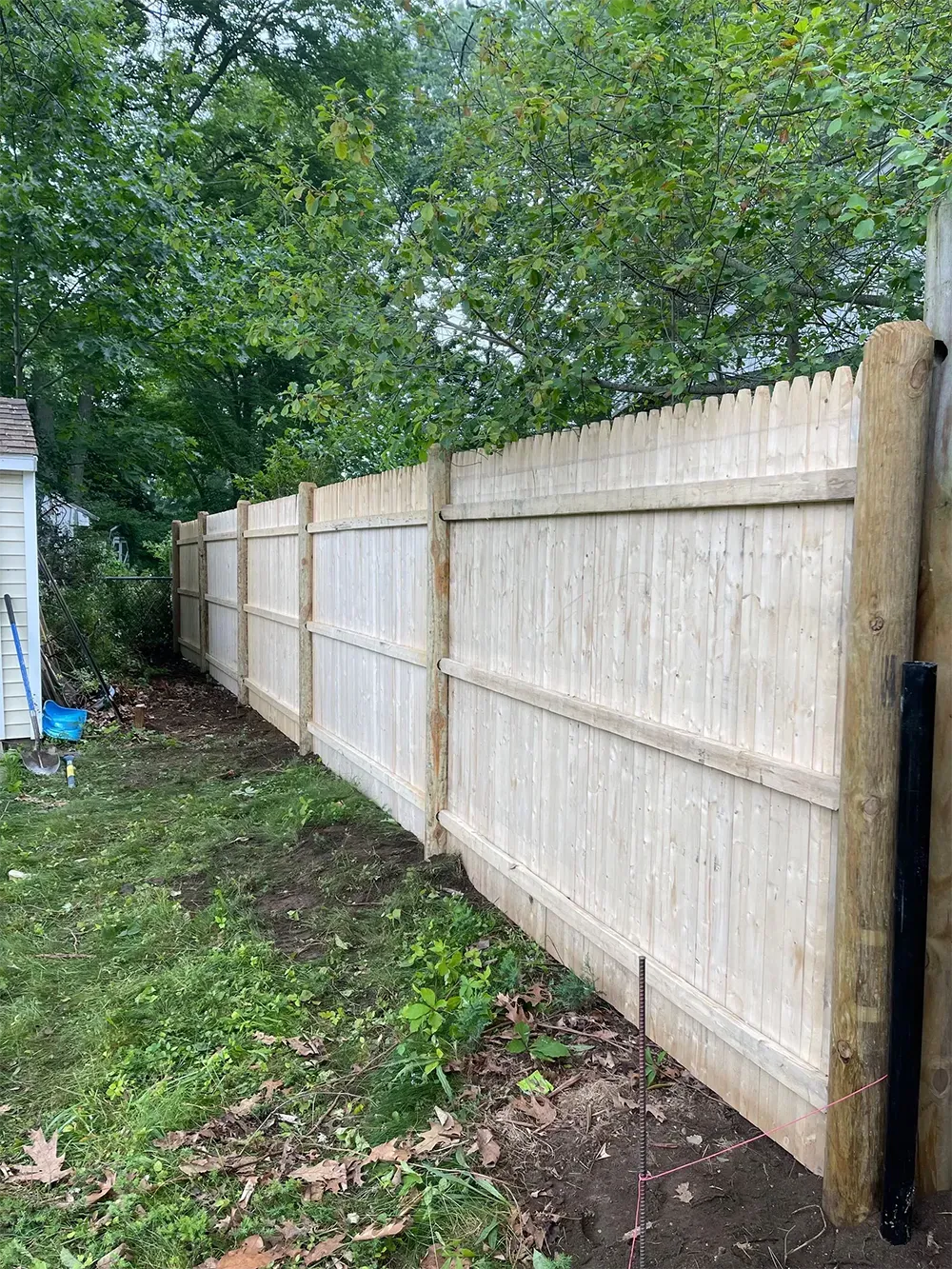 Wooden fence in a backyard, brown posts and light wood panels. Green grass and trees in background.