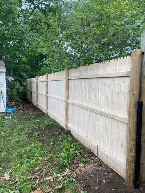 Wooden fence in a backyard, brown posts and light wood panels. Green grass and trees in background.