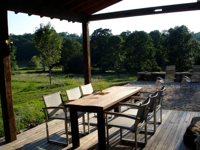 Outdoor patio with a wooden dining table and six chairs, overlooking a green field with trees in the background, under a wooden pergola.