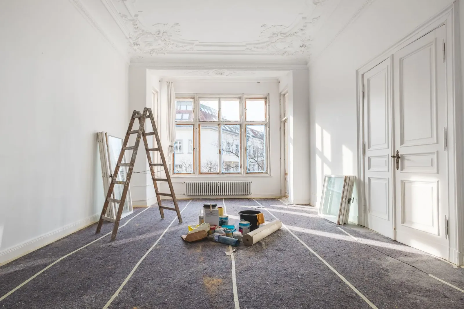 A bright room under renovation with a ladder, painting supplies, and protective floor covering.