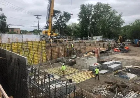 Construction site with workers, concrete structures, and heavy machinery, including a yellow crane, under cloudy skies.
