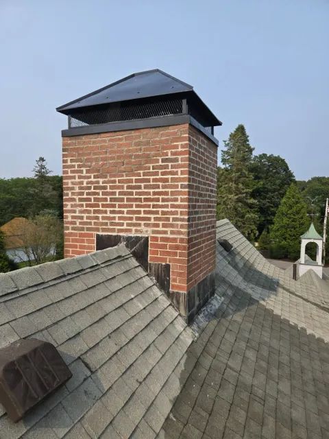 Brick chimney with a black cap on a gray shingled roof, trees in the background.