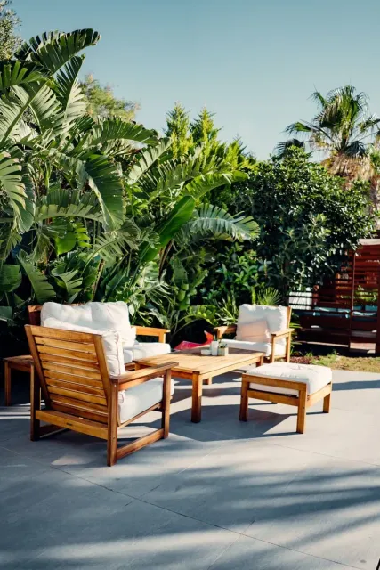 Wooden patio chairs and ottoman on a sunlit terrace surrounded by lush tropical plants