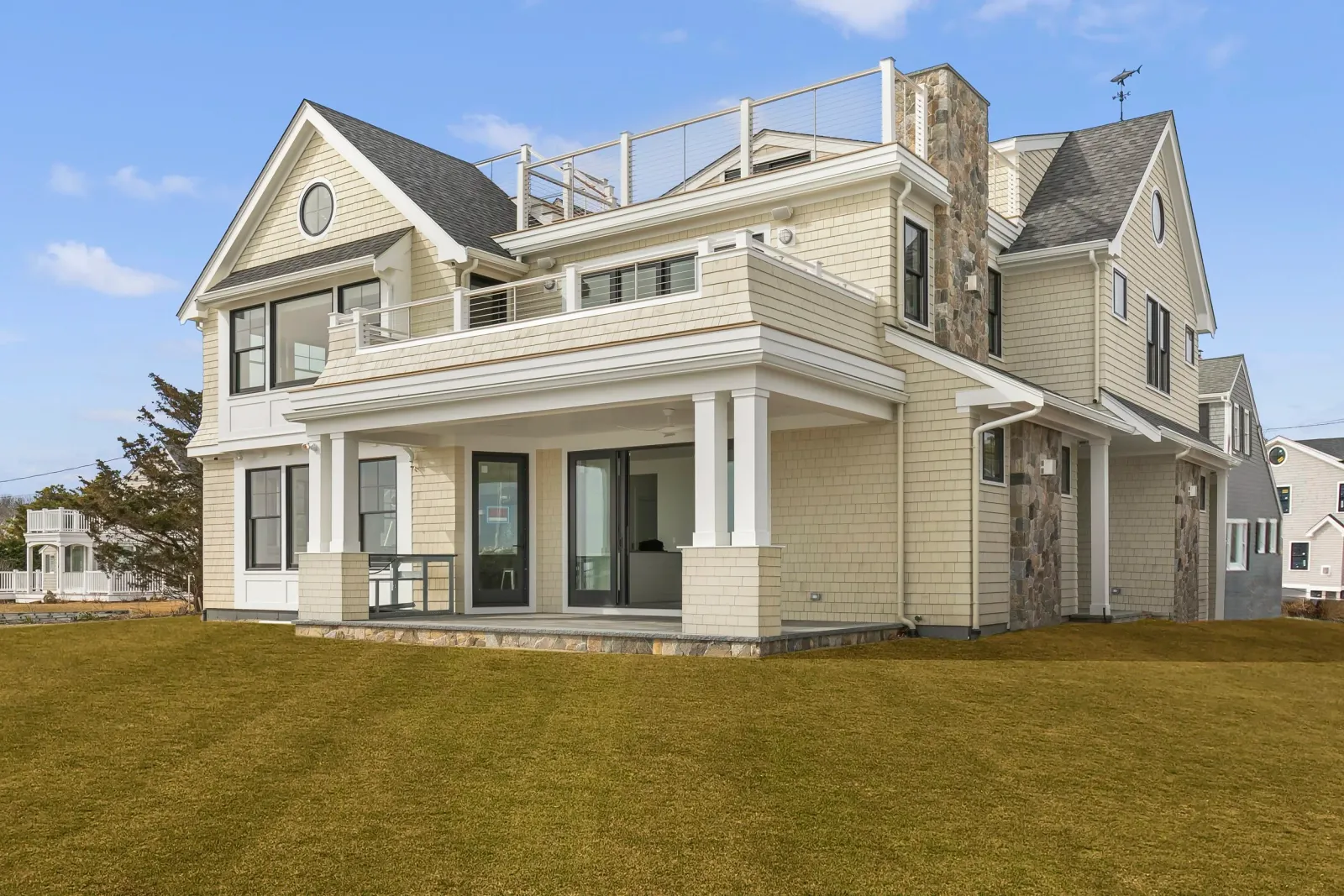 A large beige and gray multi-story house with white trim, multiple pitched roofs, a stone chimney, and patio area with glass sliding doors, on a grassy lawn under a blue sky.