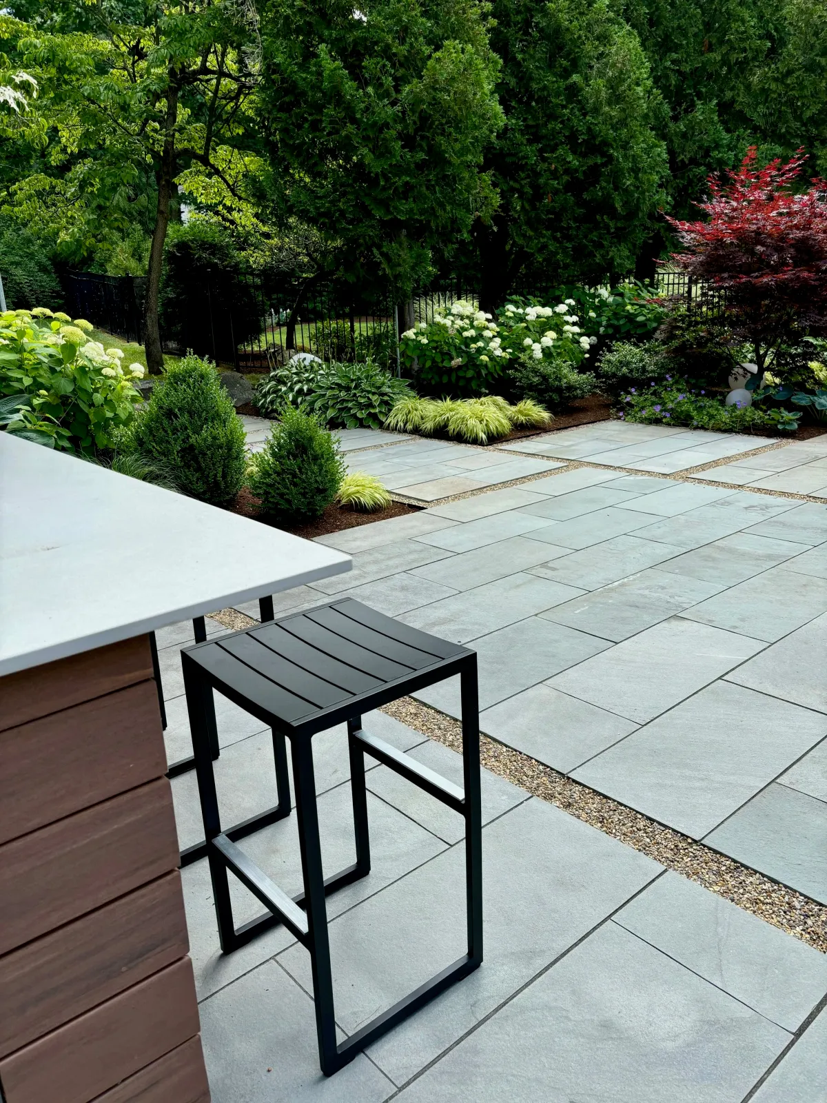 An outdoor kitchen complete with a grill station, outdoor sink and fridge and bar seating with granite patio and adjacent garden design are a highlight of this Midcentury Modern home in Andover, MA.
