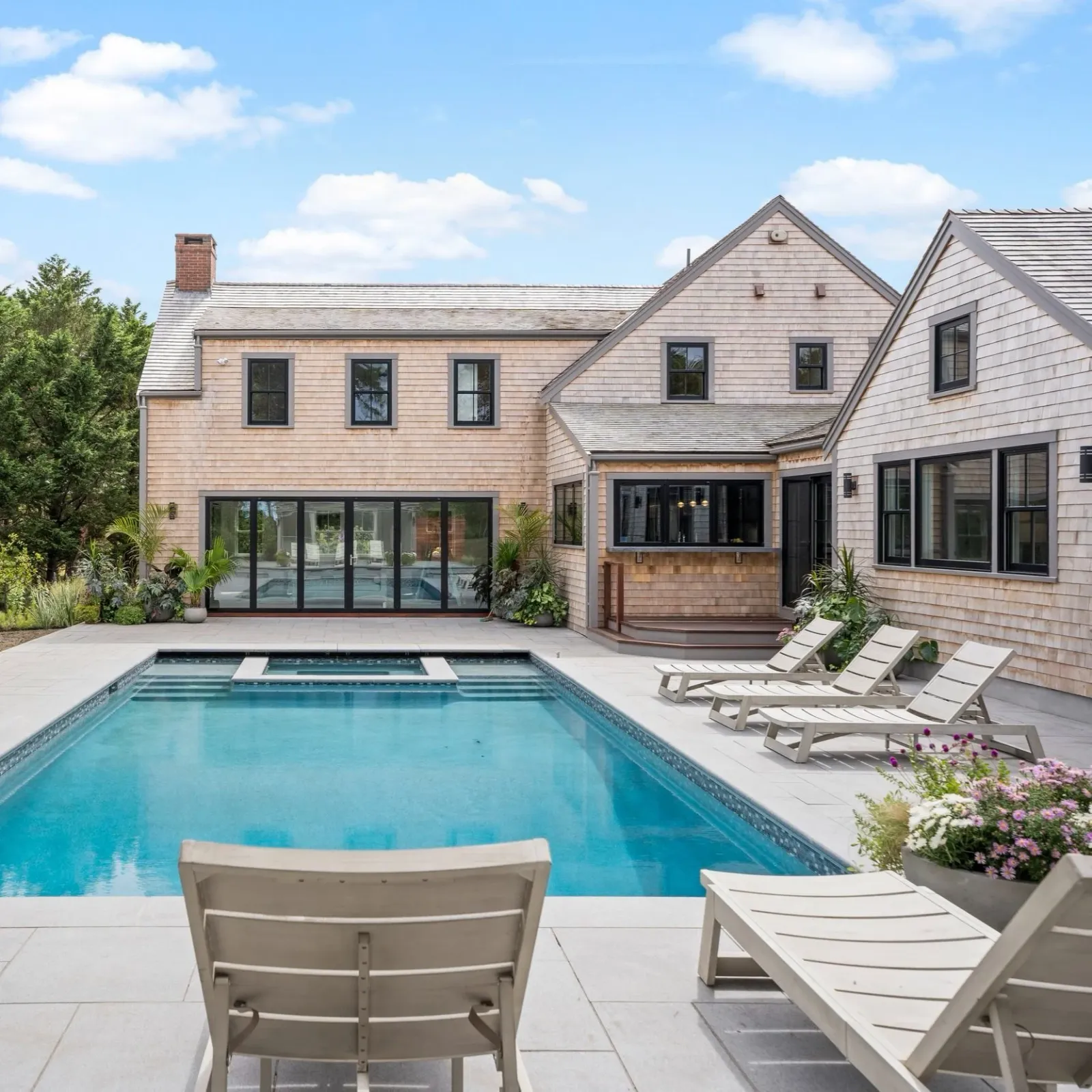 Modern house with multiple windows and wood siding, backyard with swimming pool, poolside chairs, and potted plants, under a partly cloudy sky.